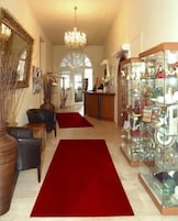 Um saguão de hotel com uma mesa de recepção, um candelabro, um tapete vermelho e uma vitrine de vidro com objetos.