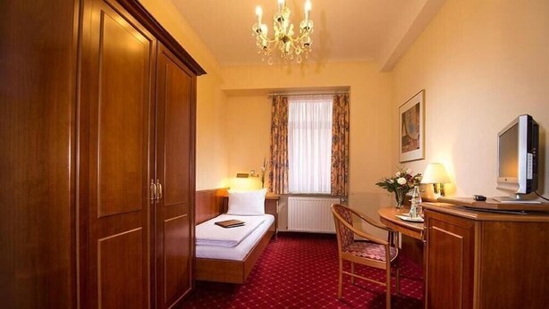 A hotel room with a bed, a desk with a chair, a television, a chandelier, and a window with curtains. - Hotel Schweizer-hof Baden Baden (Baden-Baden)