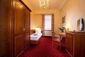 Quarto de hotel com cama, escrivaninha com cadeira, televisão, lustre e janela com cortinas.