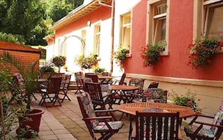 Um pátio externo com mesas e cadeiras de madeira, plantas em vasos e um edifício vermelho com janelas.