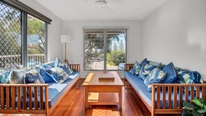 Ein Zimmer mit Holzboden, Deckenventilator, Sofagarnitur, Tisch und großen Fenstern mit Ausblick.