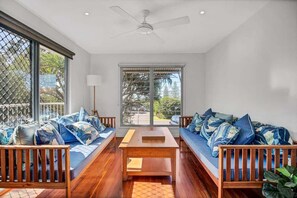 A room with a wooden floor, a ceiling fan, a sofa set, a table, and large windows with a view.