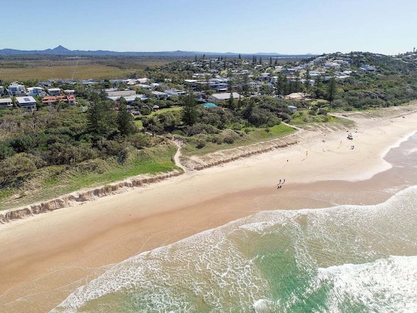 A coastal area with a sandy beach, a residential neighborhood, and a hilly landscape in the background.