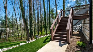 Eine Holztreppe führt zu einem Haus mit Metallgeländer, umgeben von einem Rasen und Bäumen.