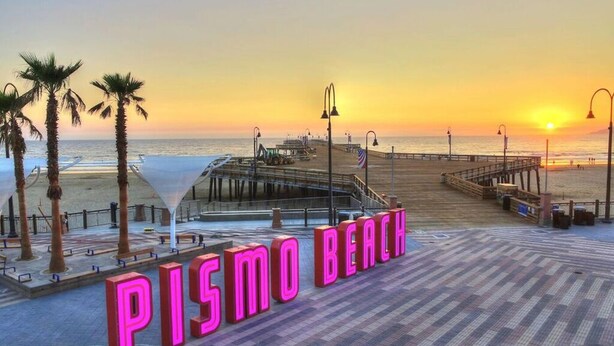 A boardwalk with the text 'PISMO BEACH' in neon lights, palm trees, and a sunset over the ocean. - Ranchito Robles, Hot Tub • Fire Pit • Wine Country Retreat (Paso Robles)