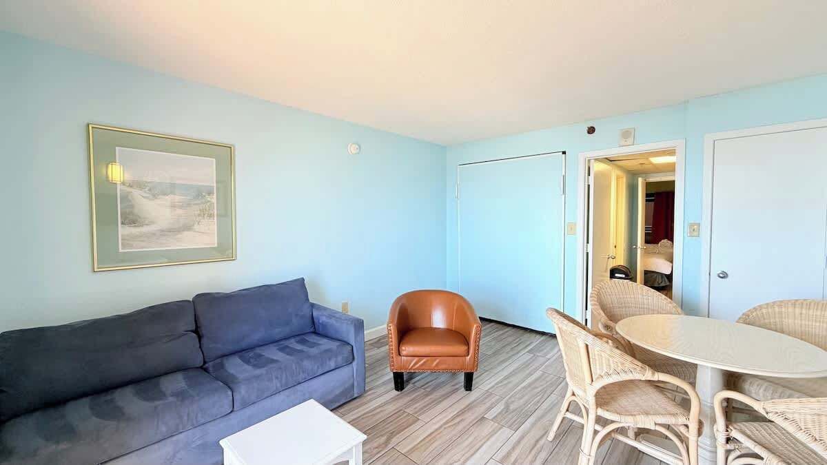 Une pièce avec un canapé bleu, un fauteuil en cuir marron, des chaises en osier, une petite table blanche et un tableau encadré accroché au mur.