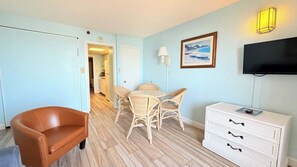 A room with a brown leather sofa, a wicker dining table with four chairs, a white dresser with drawers, a flat-screen TV mounted on the wall, and a framed picture above the TV. - Direct Oceanfront 1BR – 3rd Floor Retreat (Myrtle Beach)