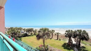 Varanda com vista para a praia, palmeiras e céu claro.