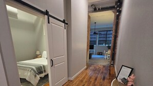 Un corridoio con pavimento in legno, un letto in una stanza a sinistra e uno scaffale bianco con oggetti incorniciati e un piccolo cesto.
