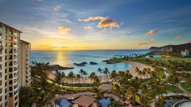 Un resort con zona de piscina, palmeras y vista al océano al atardecer.