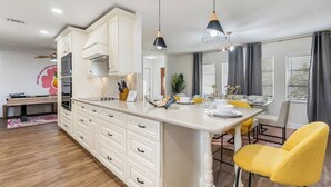 Une cuisine moderne avec des meubles blancs, un îlot central, des luminaires suspendus et un coin repas avec des chaises jaunes.