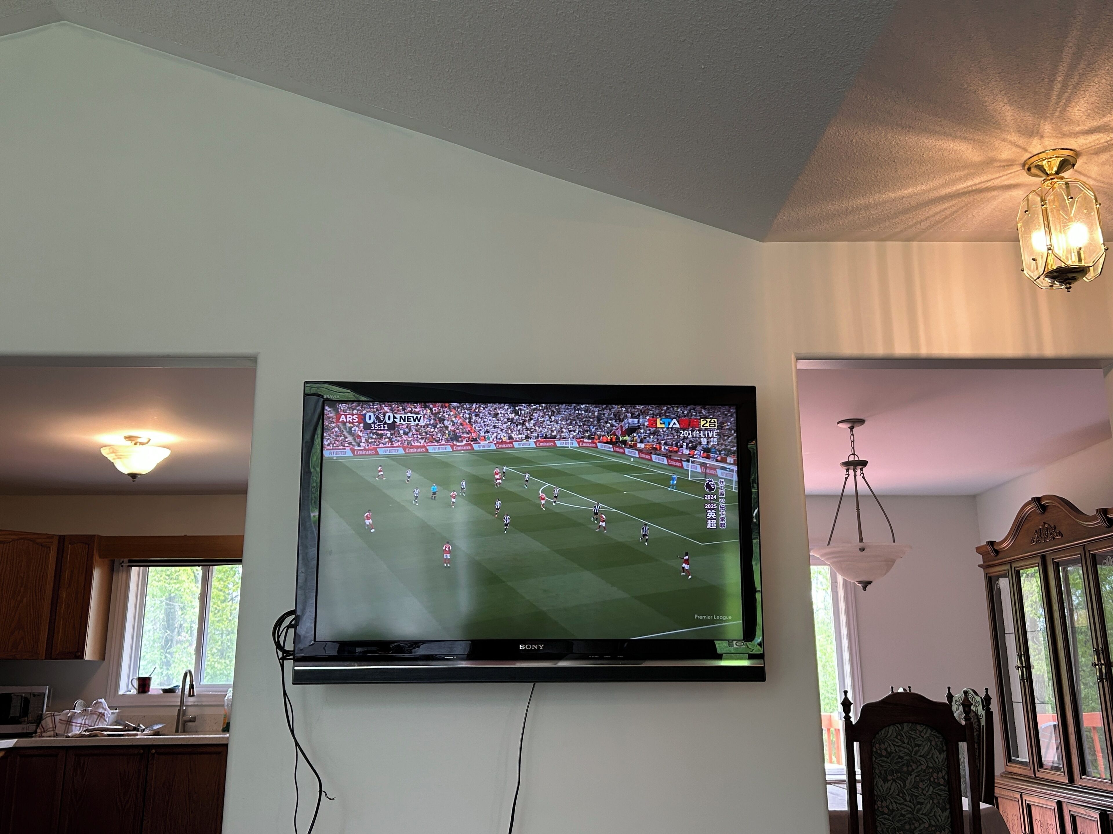 Ein an der Wand befestigter Flachbildfernseher zeigt ein Fußballspiel, darunter befindet sich eine Küchenzeile und ein Esstisch.