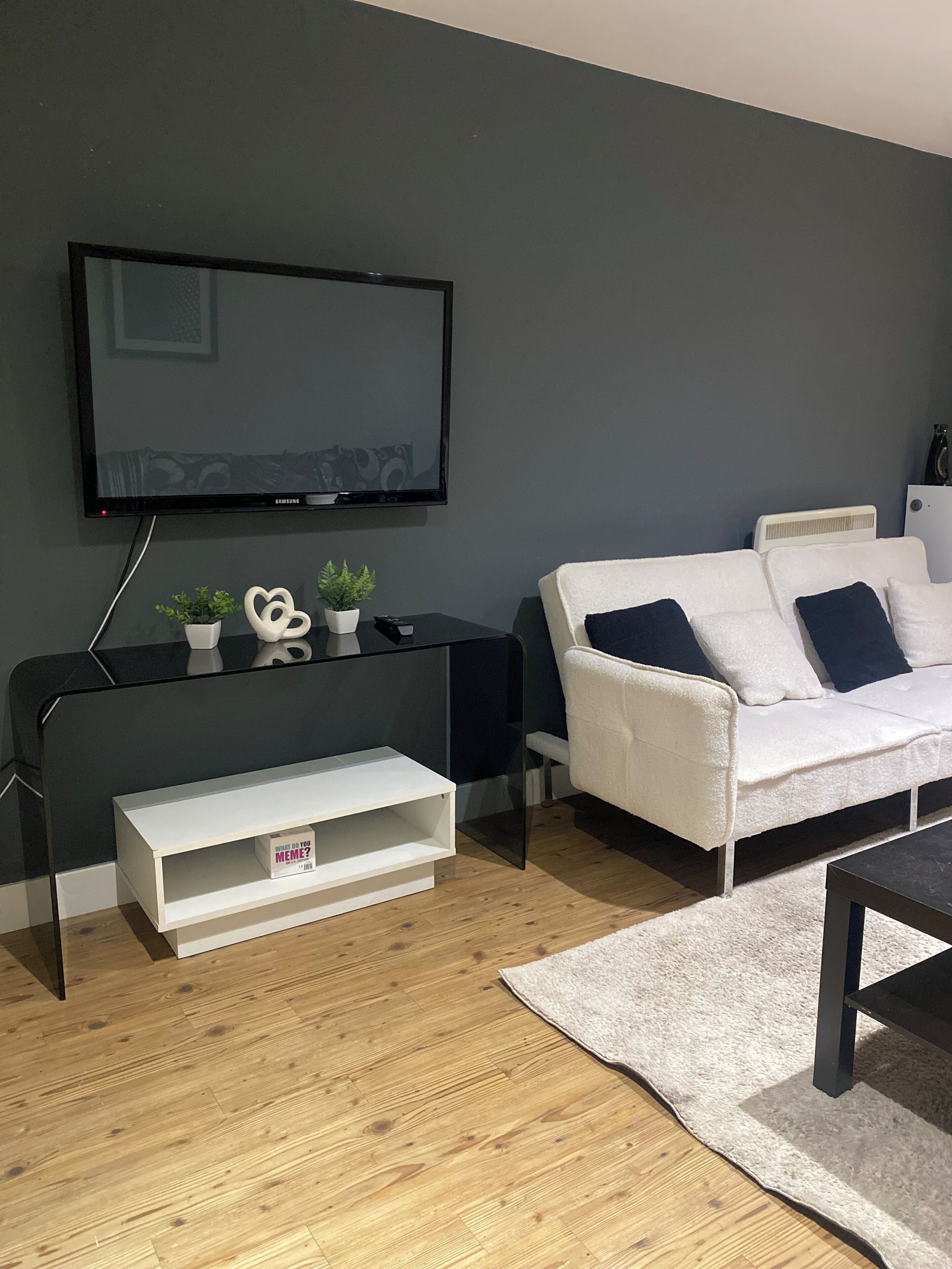 A modern living room with a flat-screen TV, a white sofa, a coffee table, and a small white shelf with decorative items.