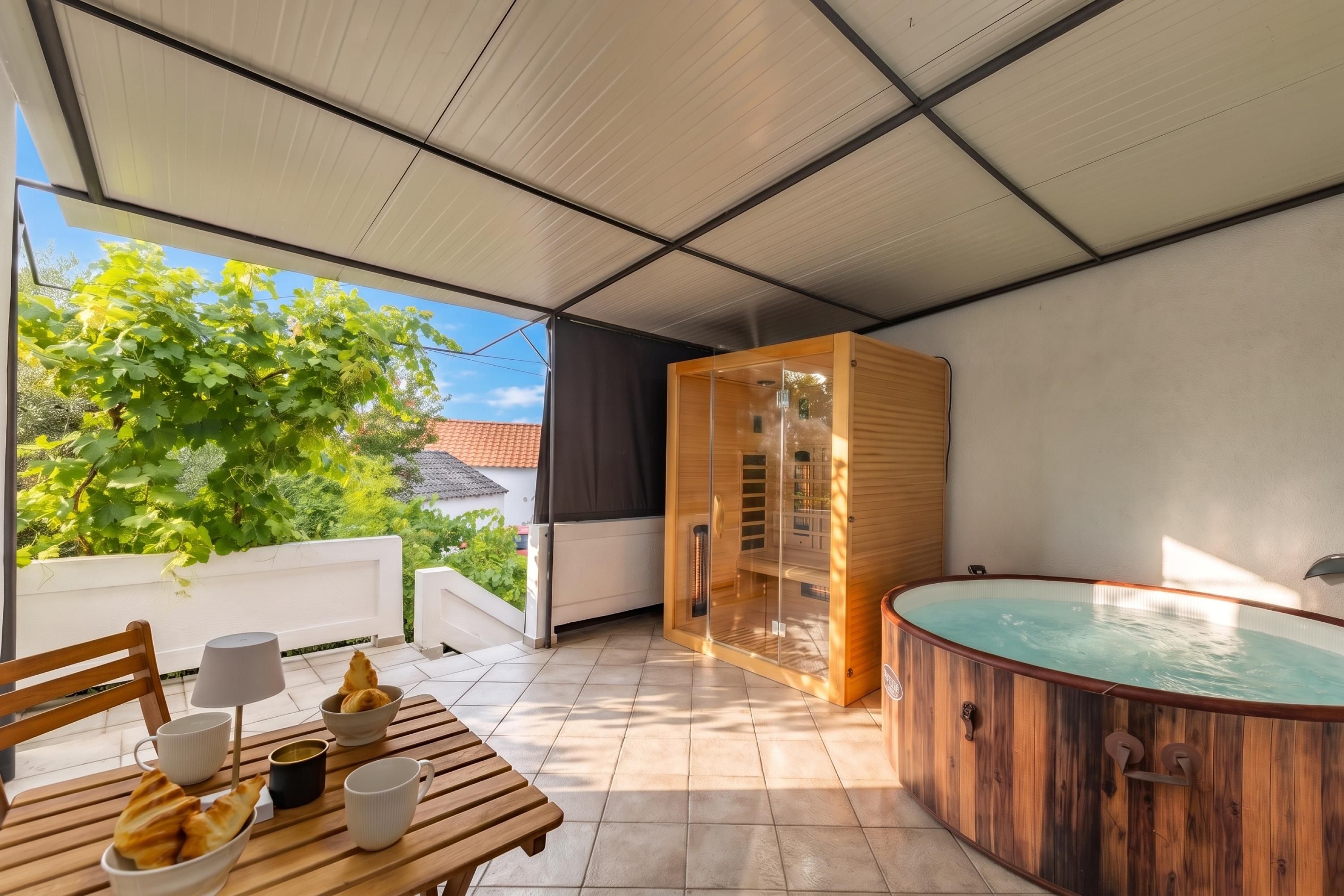 Un espacio exterior cubierto con una mesa de madera, sillas, un jacuzzi y una sauna con paredes de vidrio.