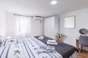 A hotel room with a bed, a bedside table, a chair, a desk, a mirror, a window with curtains, an air conditioning unit, and a wall-mounted mirror.