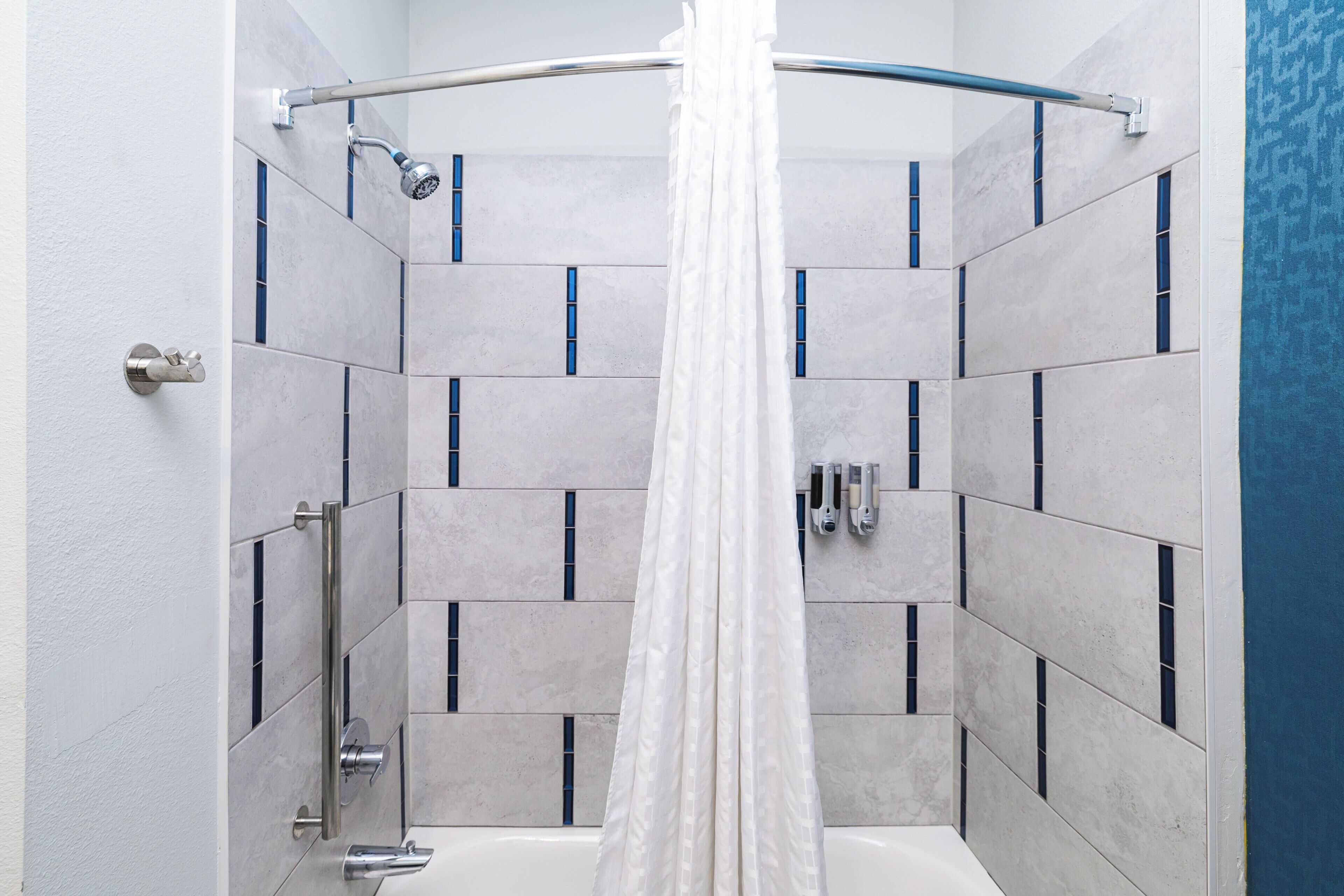 A shower with a white curtain, a handheld showerhead, and a handheld soap dispenser.