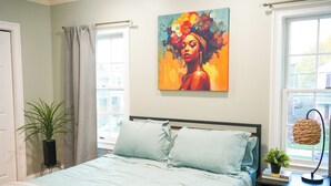 A bedroom with a bed, a painting, a potted plant, and a window.