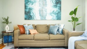 A living room with a beige sofa, a large abstract painting, a potted plant, and a small side table with a vase and books.