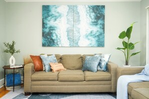 Een woonkamer met een beige bank, een groot abstract schilderij, een potplant en een klein bijzettafeltje met een vaas en boeken.