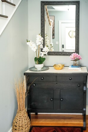 Een donkere houten dressoir met een spiegel erboven, een witte vaas met witte bloemen, een gouden schaal, en enkele boeken.