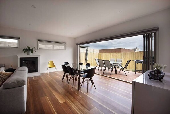 A modern living room with a fireplace, a dining area with chairs, and a large sliding glass door leading to an outdoor patio.
