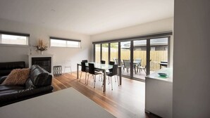 A modern living room with a fireplace, a dining area with a large table and chairs, and a view through large sliding glass doors.