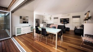 A modern kitchen with a dining area, a living room with a sofa and TV, and a sliding glass door leading to an outdoor area.