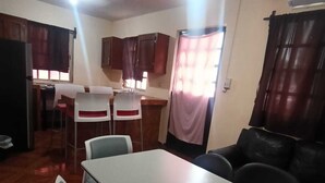 Uma sala de jantar com mesa e cadeiras, cozinha ao fundo e um ventilador de teto.