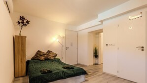 A bedroom with a bed, a wooden wardrobe, a nightstand, a wall-mounted lamp, and a door leading to another room.
