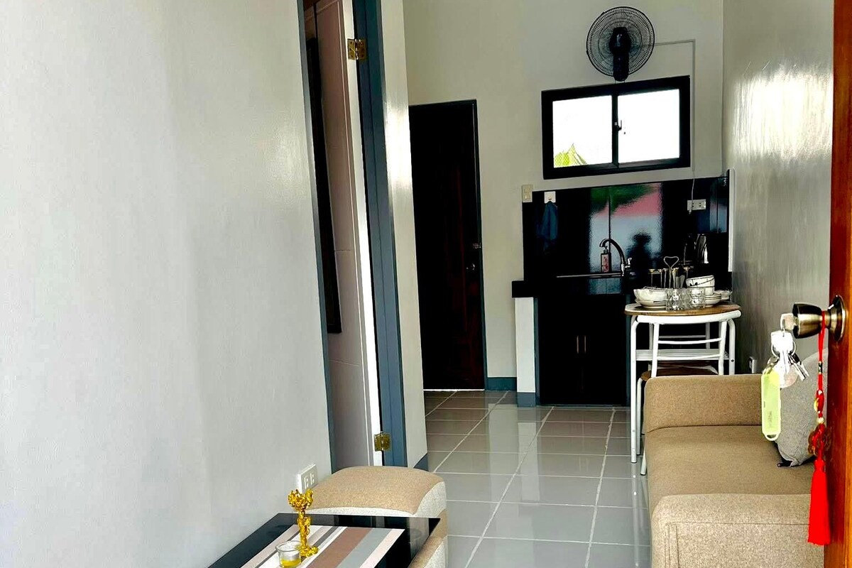 A modern living room with a sofa, ottoman, and a small table with a decorative item. A television is mounted on the wall, and a fan is visible in the background.