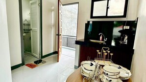 Ein Essbereich mit einem runden Holztisch, gedeckt für vier Personen, einem schwarzen Kühlschrank, einem Fenster mit Jalousien und einer Tür, die auf einen Balkon führt.