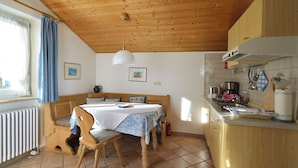 Ett mysigt rum med ett träpanelat tak, ett matbord med stolar, en bänk, en köksdel med en spis och ett fönster med gardiner.
