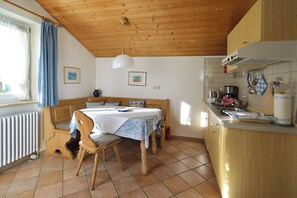 A cozy room with a wooden ceiling, a dining table with chairs, a bench, a kitchen area with a stove, and a window with curtains. - Apartment 50sqm, with balcony (Seebruck)