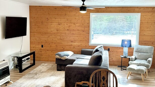 A modern living room with a wooden wall, a ceiling fan, a flat-screen TV, a sofa, a dining table set for four, and a window with a view of trees.