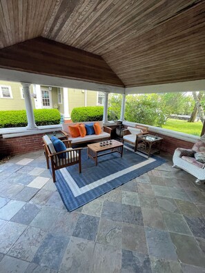 Un patio cubierto con piso de piedra, techo de madera y zona de estar con sofá y sillones.