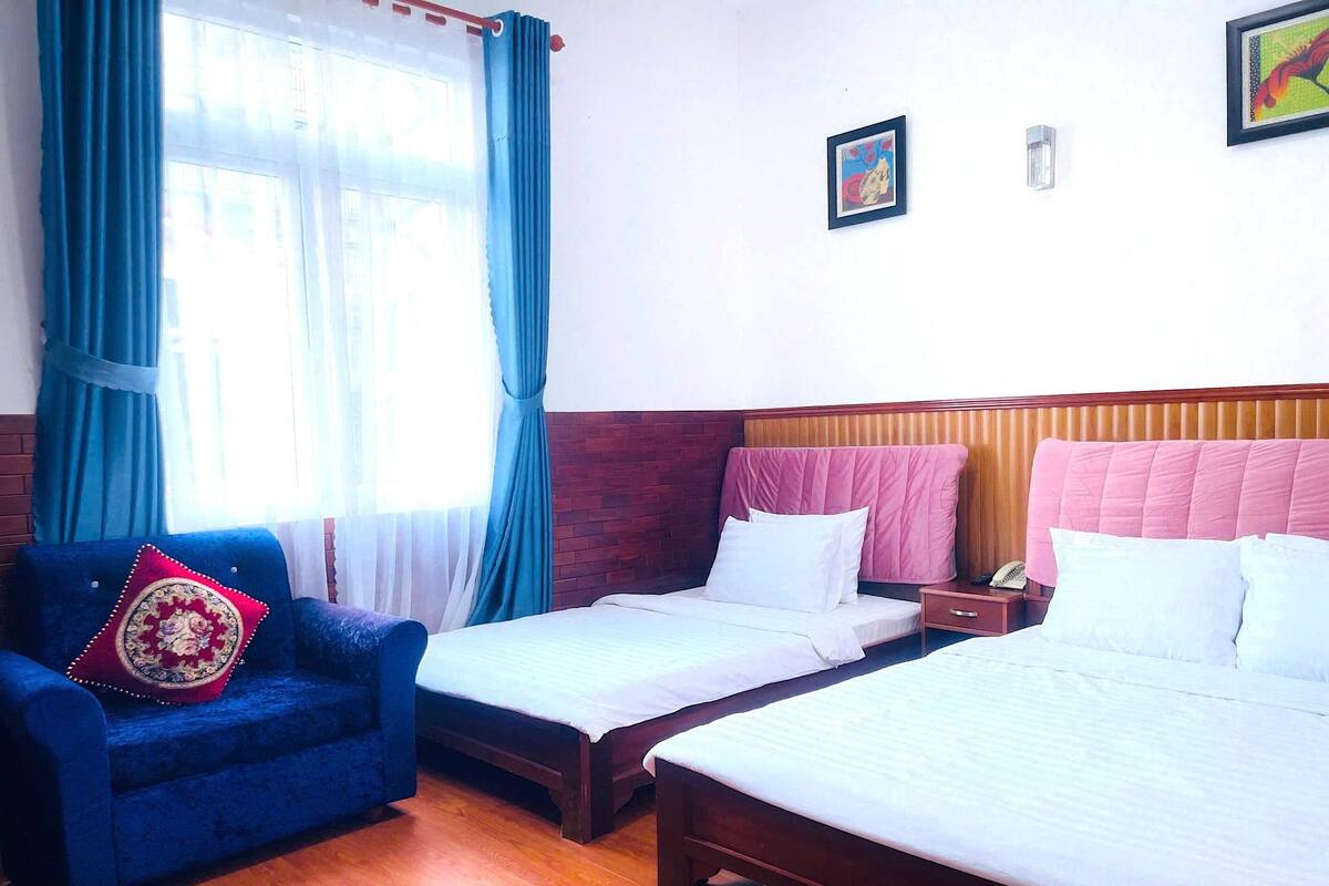 Une chambre d’hôtel avec deux lits simples, un fauteuil bleu agrémenté d’un coussin décoratif, une tête de lit en bois et une fenêtre avec des rideaux bleus.