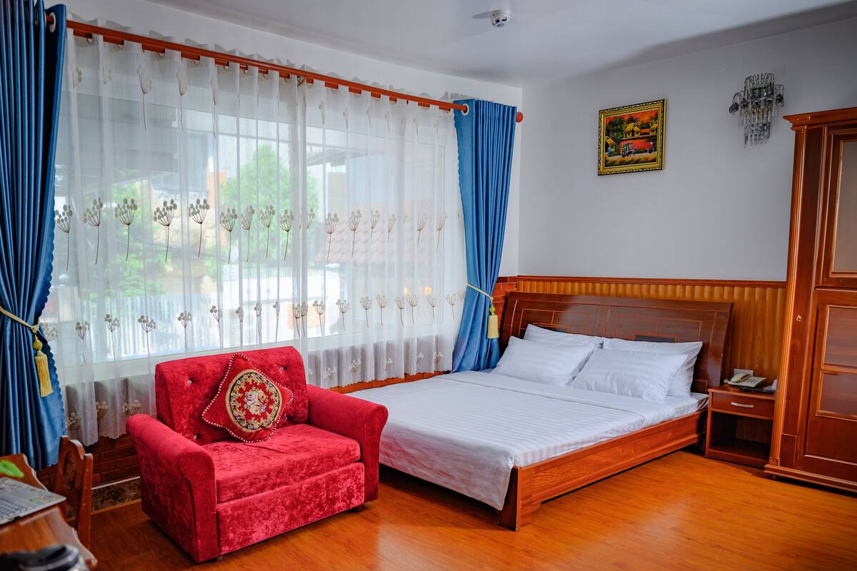 Une chambre à coucher avec un lit, une chaise rouge, une armoire en bois et une fenêtre avec des rideaux.