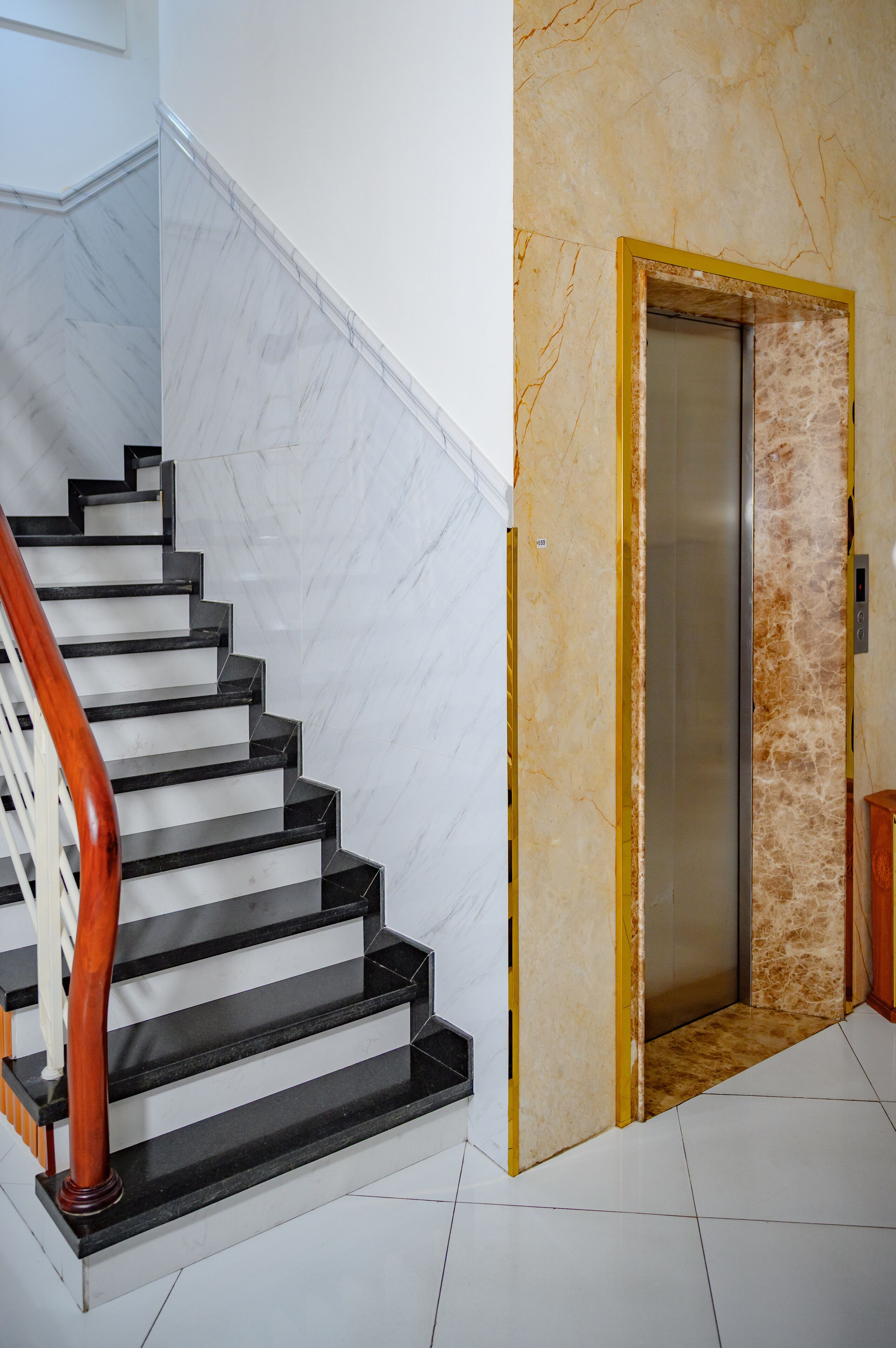 A staircase with black steps and a wooden handrail, leading to an elevator with a marble wall.