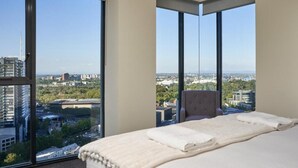 A hotel room with a large bed, a chair, and a view of a cityscape through floor-to-ceiling windows. - Exclusive Stays - The Sentinel  (Southbank)