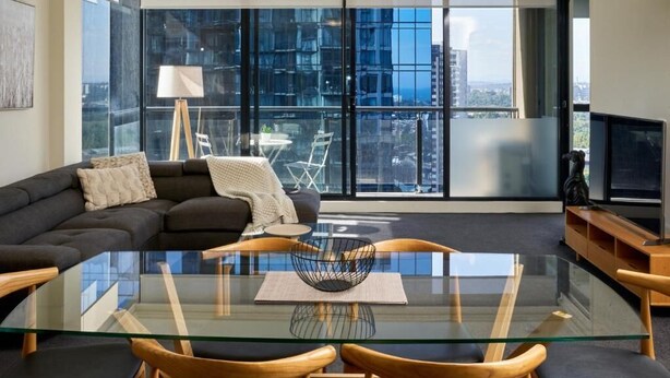 A modern living room with a glass-top dining table, a dark sofa, and a large window with a city view. - Exclusive Stays - The Sentinel  (Southbank)
