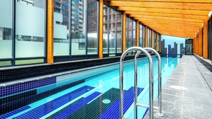 A modern indoor swimming pool with clear blue water, wooden beams, and large windows offering views of the city. - Exclusive Stays - The Sentinel  (Southbank)