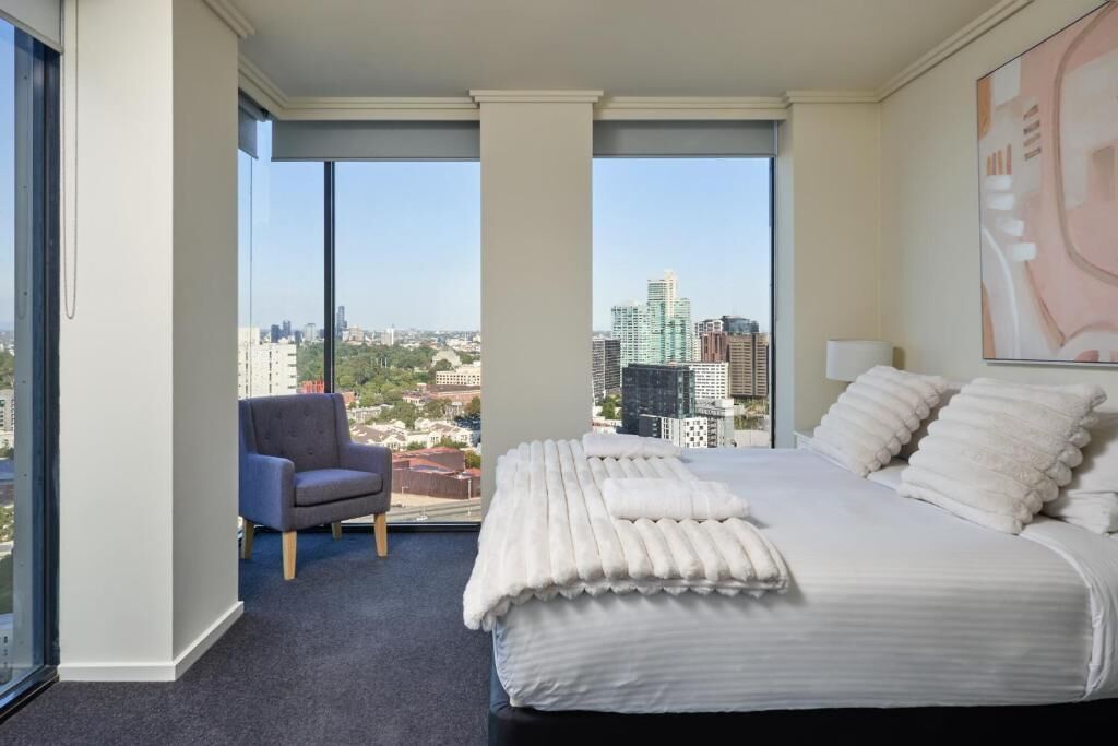 A hotel room with a large bed, a chair, and a view of the cityscape through large windows.