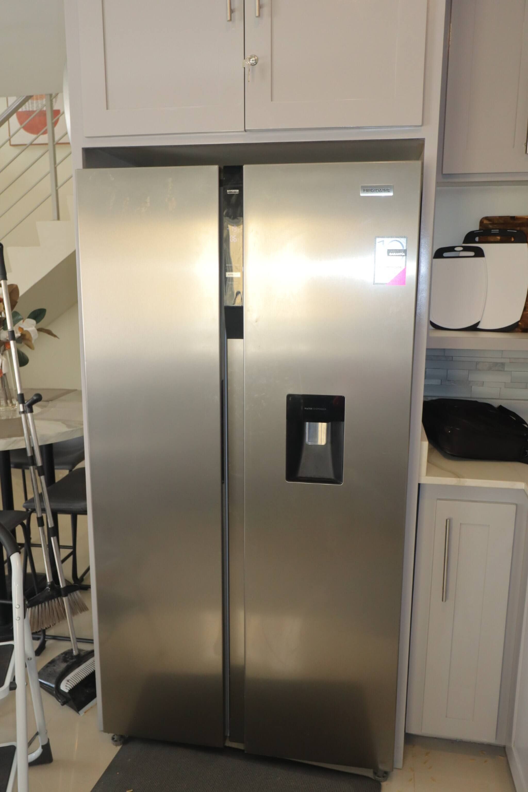 Un refrigerador de acero inoxidable con dispensador de agua, ubicado en una cocina con gabinetes blancos y salpicadero de azulejos.