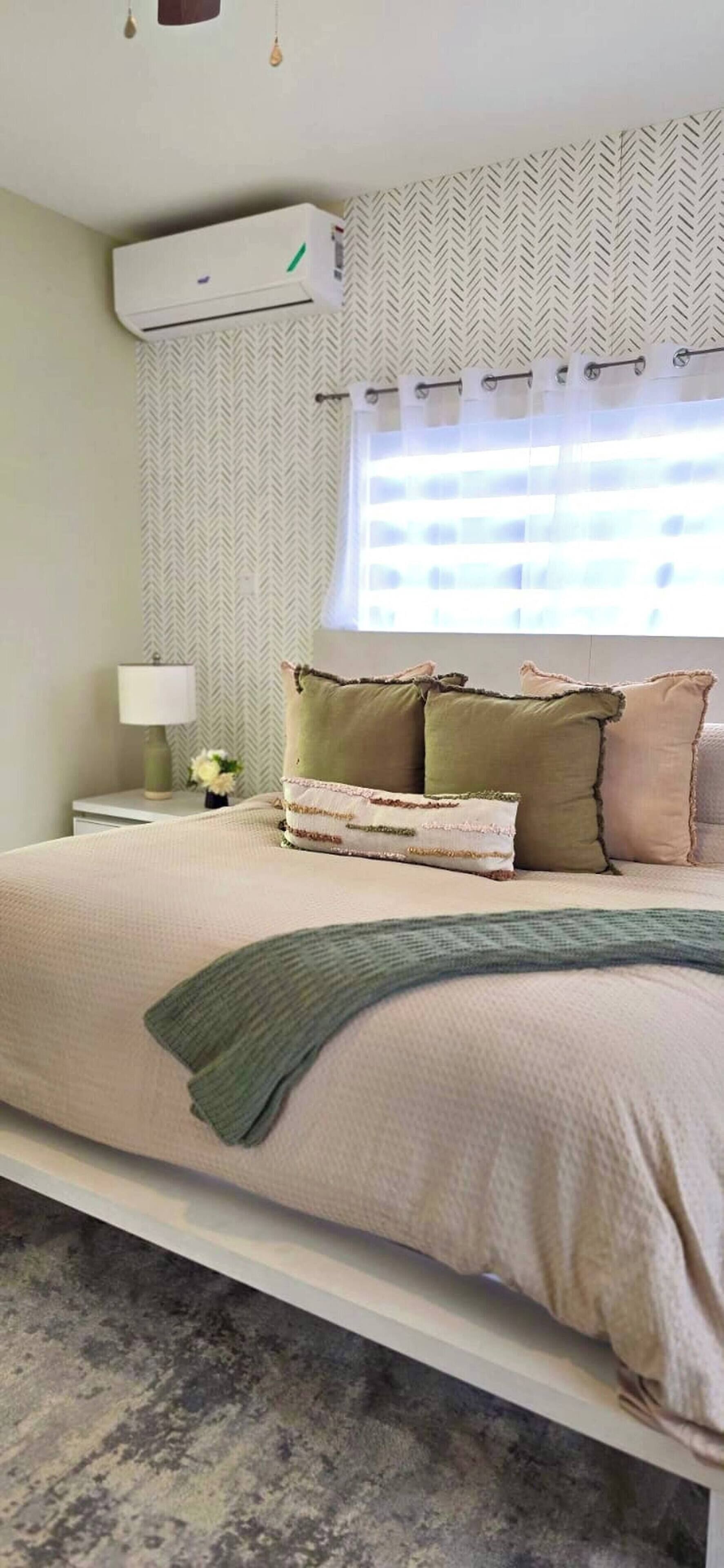A bedroom with a bed, pillows, a blanket, an air conditioner, a window with curtains, and a wall with a herringbone pattern.