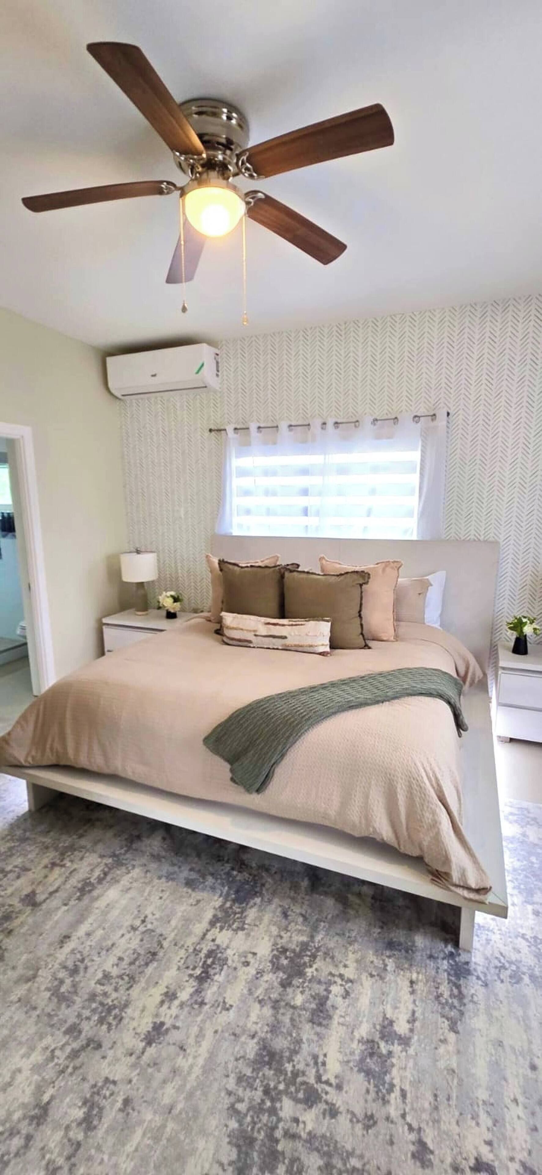 A bedroom with a bed, a ceiling fan, an air conditioning unit, a window with curtains, and a nightstand with a lamp.