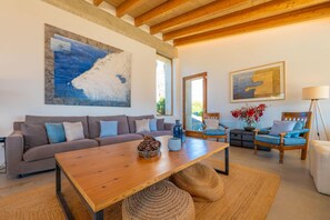 Un salon moderne avec une table en bois, un canapé et deux fauteuils. Des tableaux ornent les murs et une lampe se trouve sur la droite.