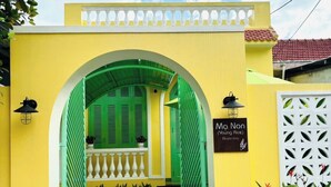 Un bâtiment jaune avec une entrée en arc vert, une enseigne indiquant « Ma Non (Young Rice) – hébergement chez l’habitant », et une clôture blanche décorative.