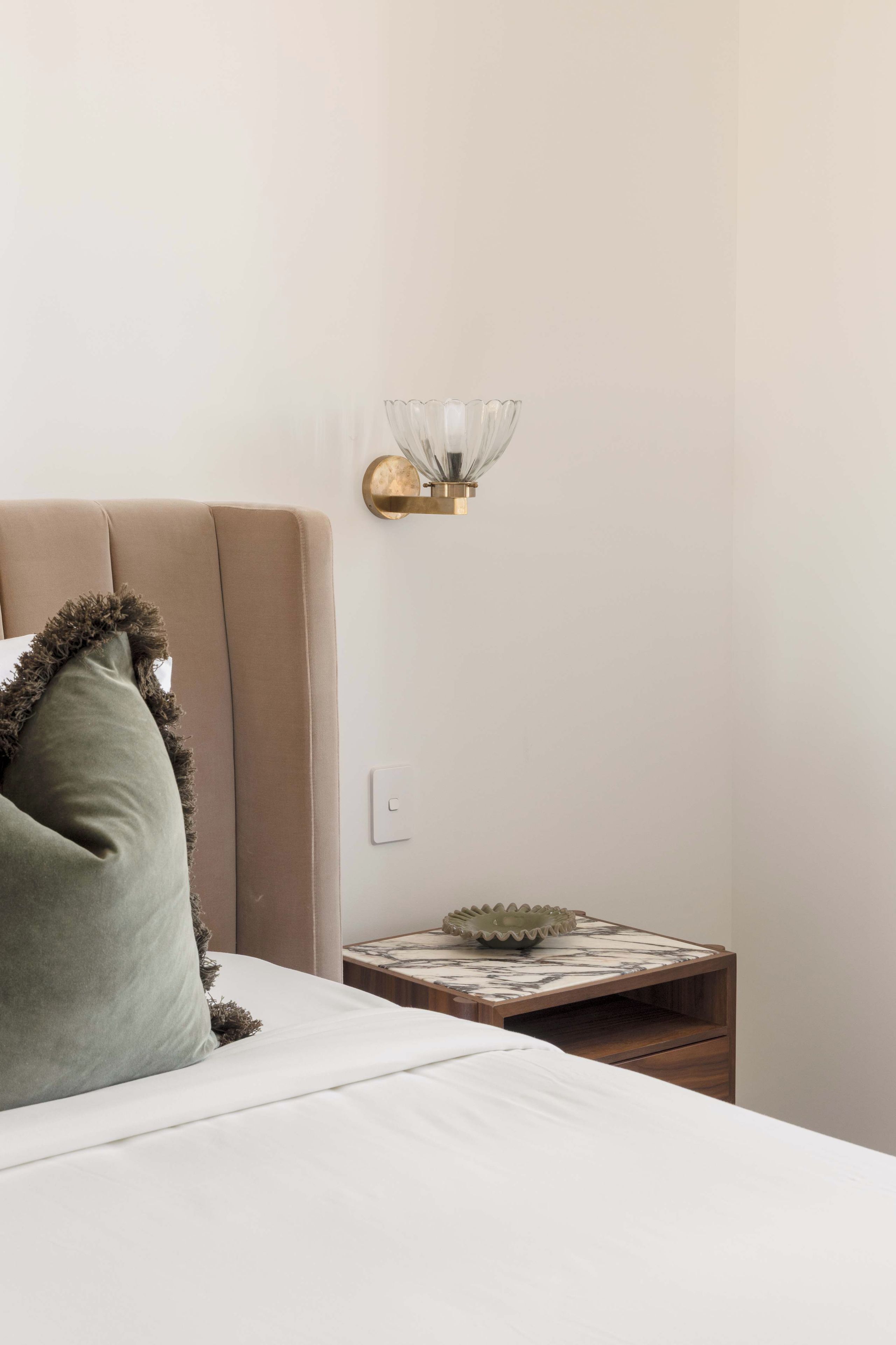 Une chambre à coucher avec un lit, une table de chevet, une lampe fixée au mur et une petite table sur laquelle se trouve un bol.