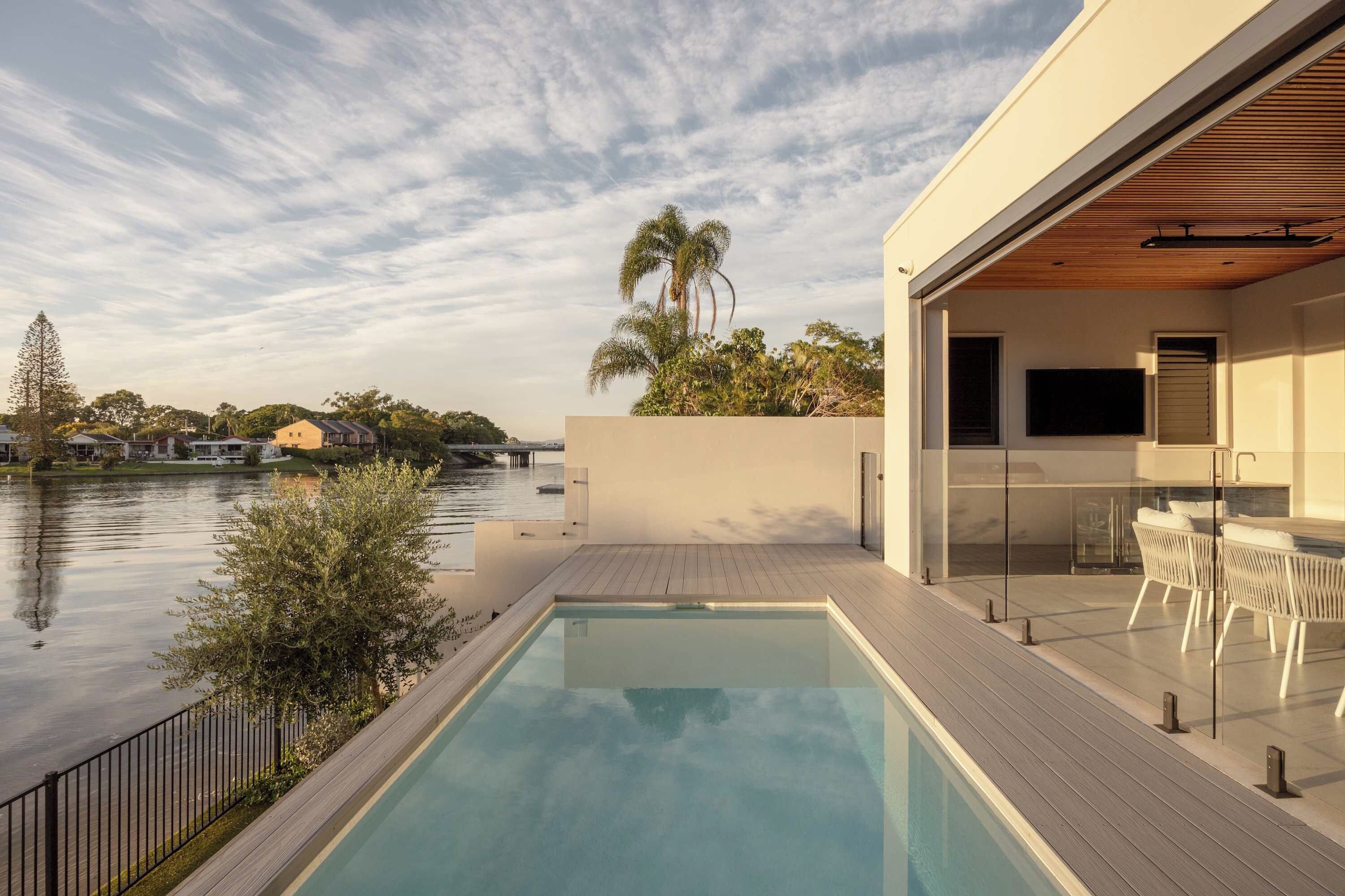 A modern outdoor pool area with a view of a river, a wooden deck, and a glass-enclosed dining area.