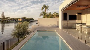 Zona moderna de piscina al aire libre con vista a un río, una terraza de madera y un área de comedor con paredes de vidrio.